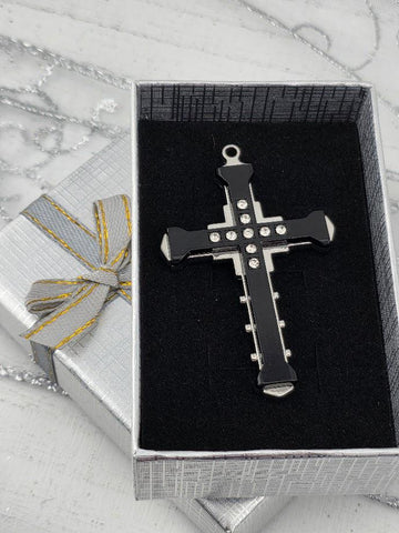 A black cross pendant with silver accents is displayed in a silver gift box with a yellow ribbon bow.