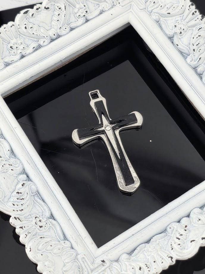 A silver cross pendant is displayed within a white ornate frame against a black background.