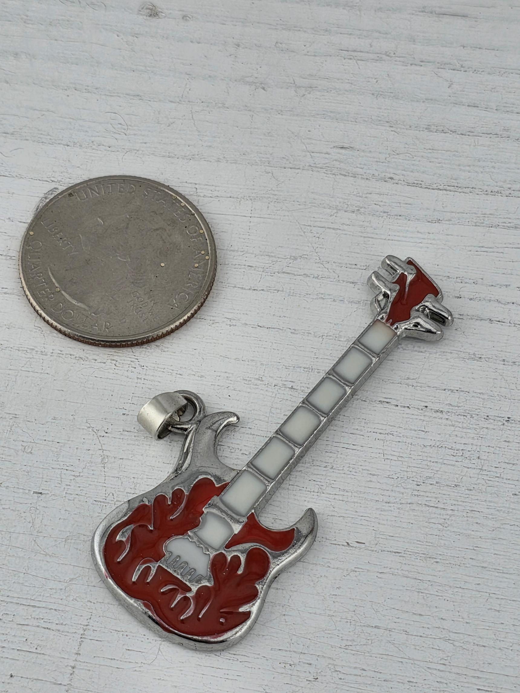 A small silver-colored guitar charm is placed next to a United States quarter coin on a white wooden surface.