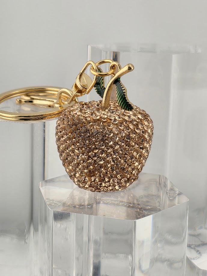 A gold-colored keychain in the shape of an apple, adorned with small rhinestones or crystals, is displayed on a clear glass stand.