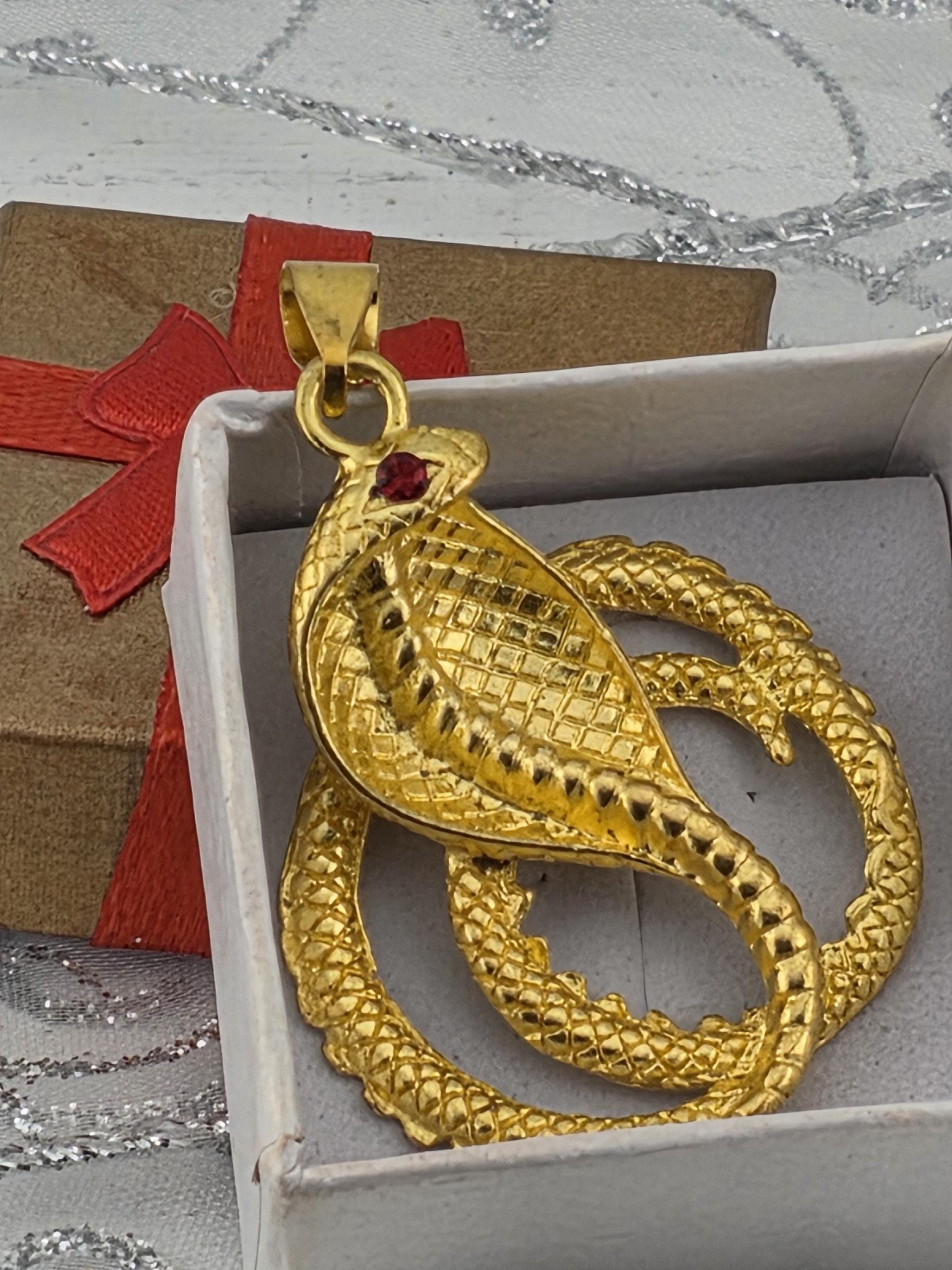 A gold snake pendant with a red gemstone is displayed in a white box, surrounded by a red ribbon and a brown gift box.