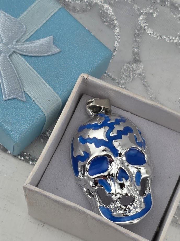 A silver and blue pendant in the shape of a skull is displayed in a white box, with a blue gift box and silver ribbon visible in the background.