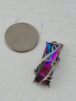 a small, colorful pendant or charm attached to a silver chain, placed next to a silver coin on a white surface.