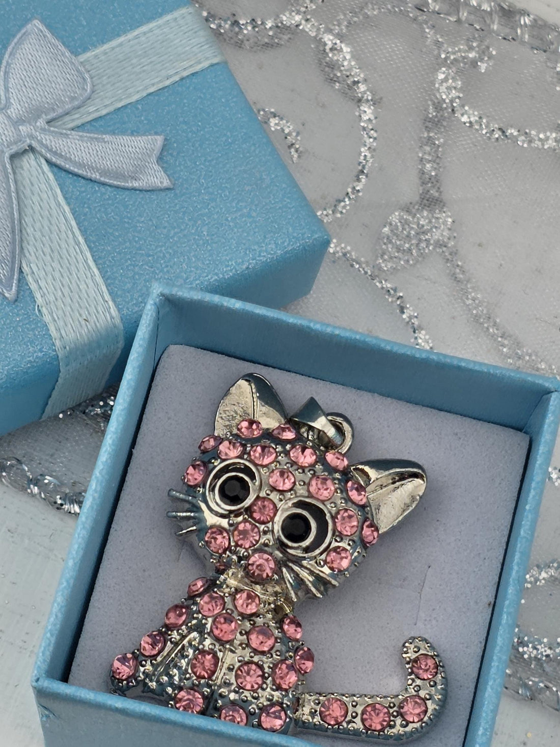 A decorative brooch in the shape of a cat, adorned with pink gemstones, is displayed in a blue gift box with a white ribbon bow.