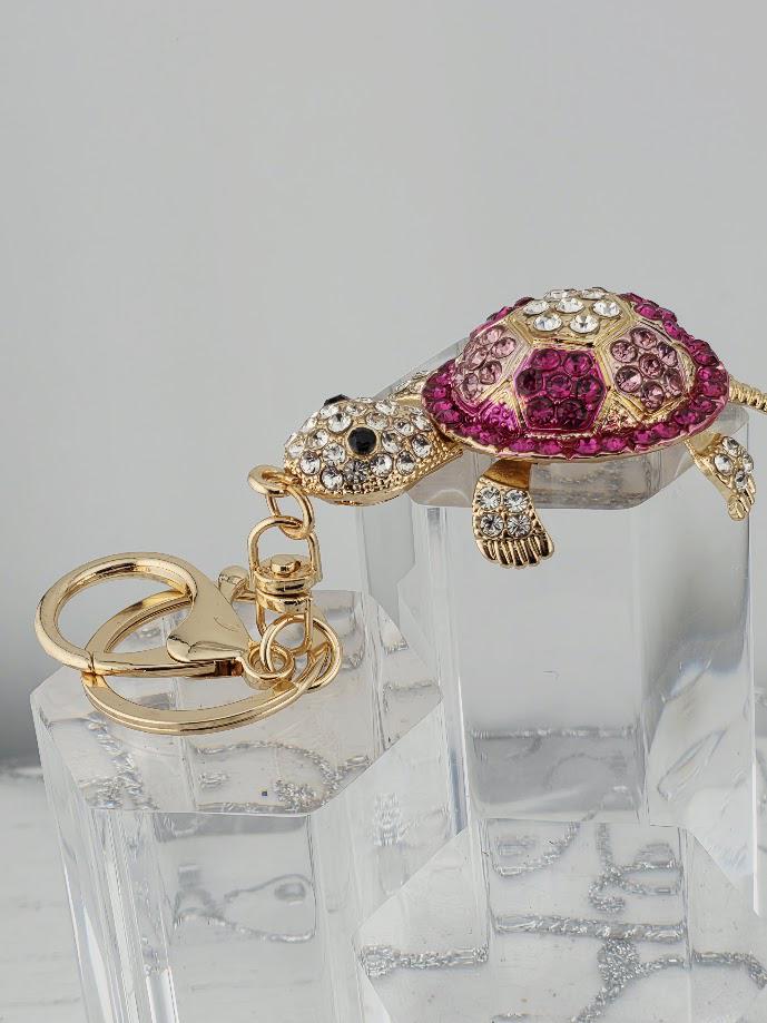 A keychain in the shape of a turtle, adorned with rhinestones and gold accents, is displayed on a clear stand.