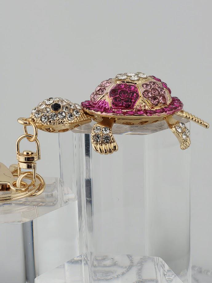A decorative keychain in the shape of a turtle, with a pink and gold color scheme and sparkling rhinestones or crystals adorning it.