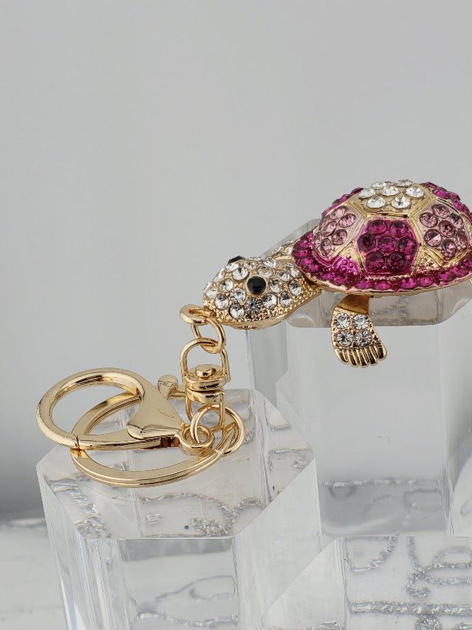 A keychain with a gold-colored charm in the shape of a turtle, adorned with pink and purple gemstones.