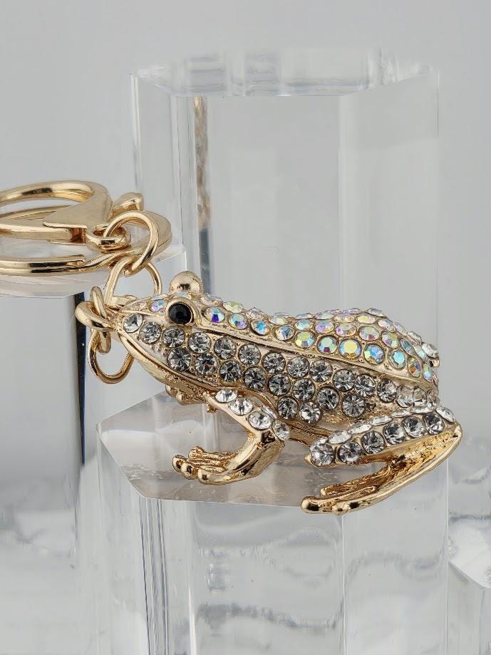 A gold-colored keychain in the shape of a frog, adorned with rhinestones or crystals, is displayed on a clear stand.
