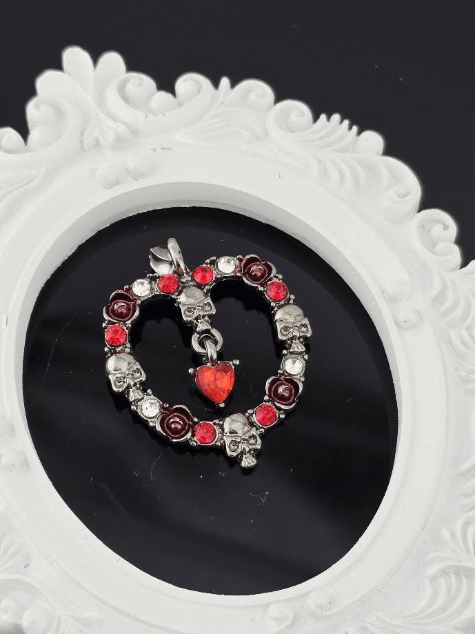A silver necklace with red gemstones and skull-shaped charms is displayed in a white box, with a brown gift box and a red ribbon visible in the background.