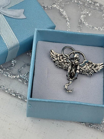 A silver-colored pendant in the shape of a winged figure is displayed in a blue gift box, with a silver ribbon tied around the box.
