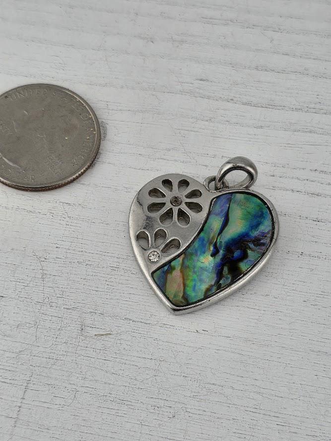 A silver heart-shaped pendant with a colorful, abstract design is shown next to a US quarter coin on a white wooden surface.