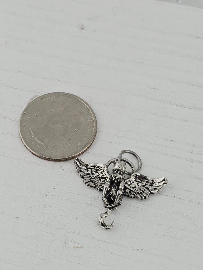 A silver-colored charm in the shape of a winged angel with a crescent moon is placed next to a United States quarter on a white surface.