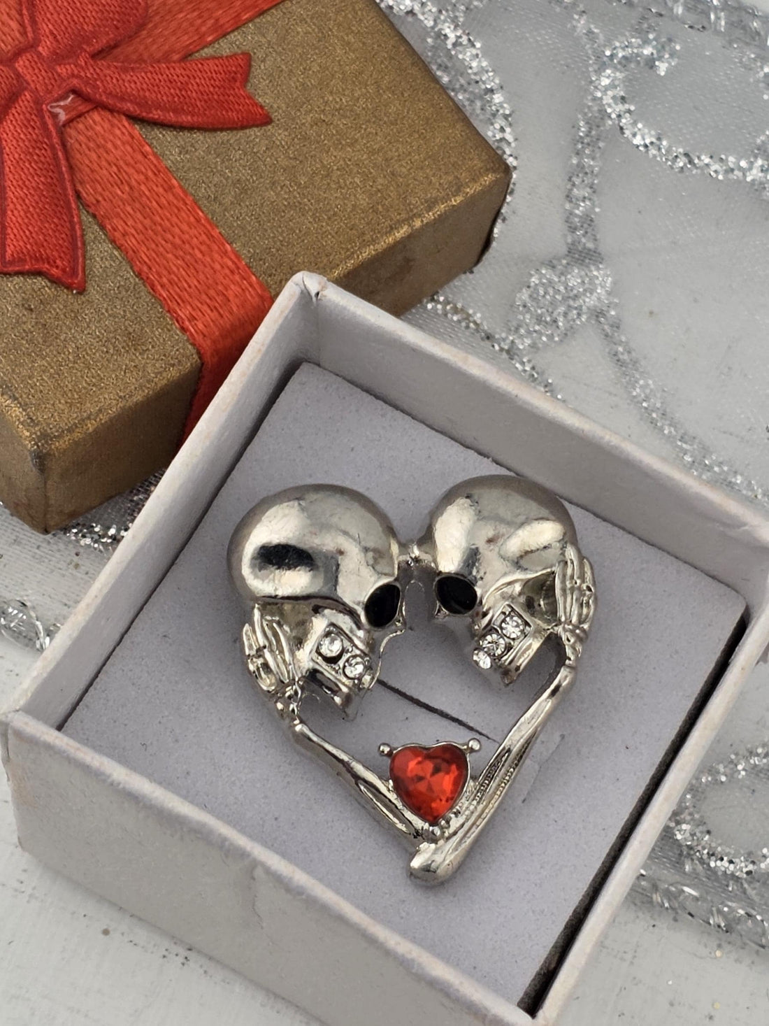 A silver brooch with two skull-shaped figures holding a red heart-shaped object is displayed in a white box with a gold ribbon.