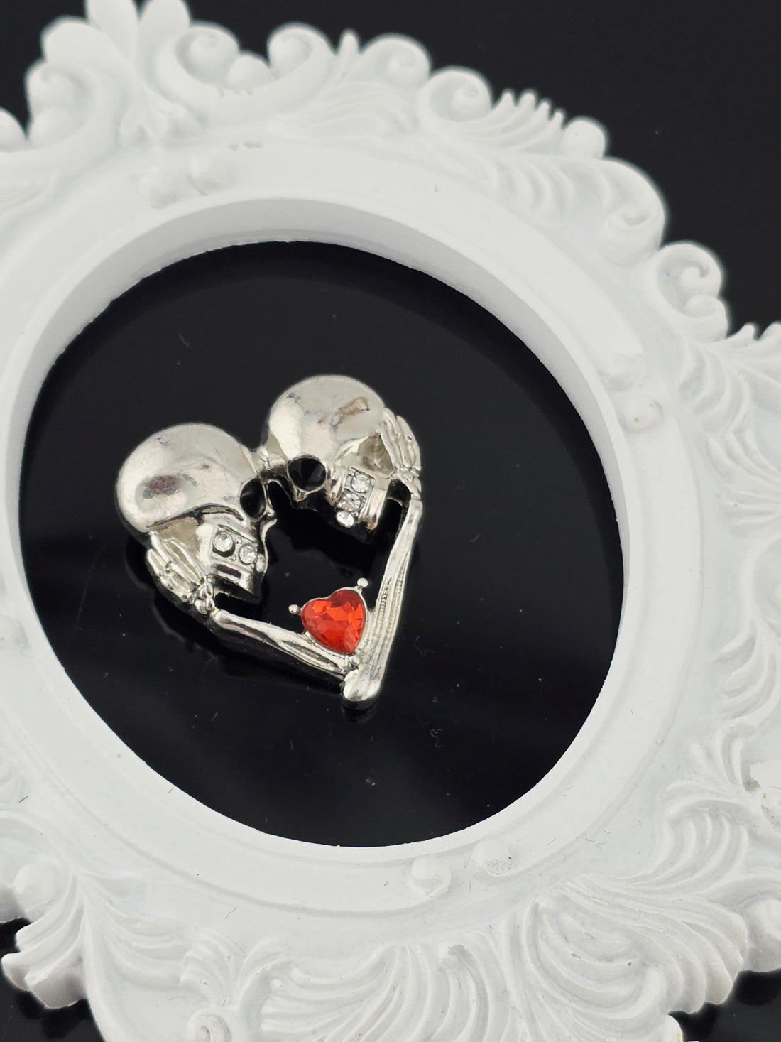 A silver brooch with two skull-shaped figures holding a red heart-shaped object is displayed in a white box with a gold ribbon.