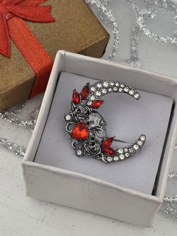 A silver brooch with a crescent moon design and red gemstones is displayed in a white box, with a brown gift box with a red ribbon visible in the background.