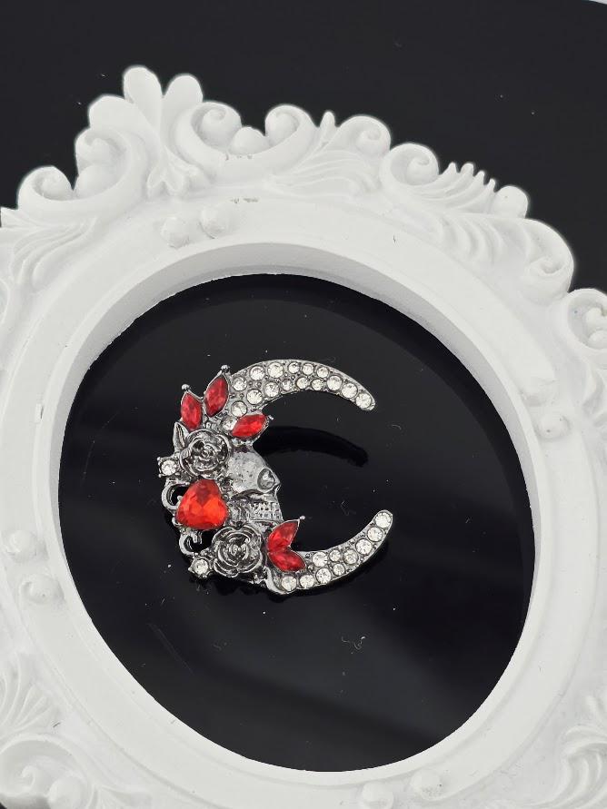 A decorative brooch in the shape of a crescent moon, adorned with red gemstones and intricate floral designs.