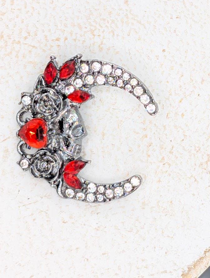A decorative brooch in the shape of a crescent moon, adorned with red gemstones and a skull design.