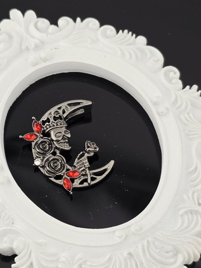 A silver brooch with a crescent moon design and red gemstones is displayed against a black background, framed by an ornate white frame.