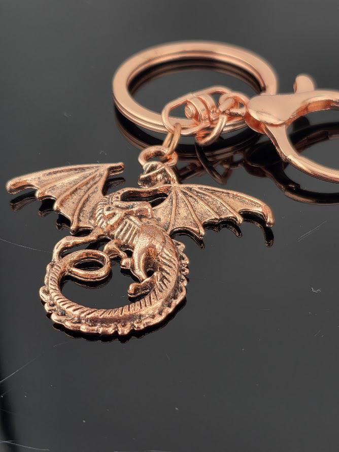 A close-up image of a copper-colored dragon keychain with wings and a tail, resting on a dark surface.
