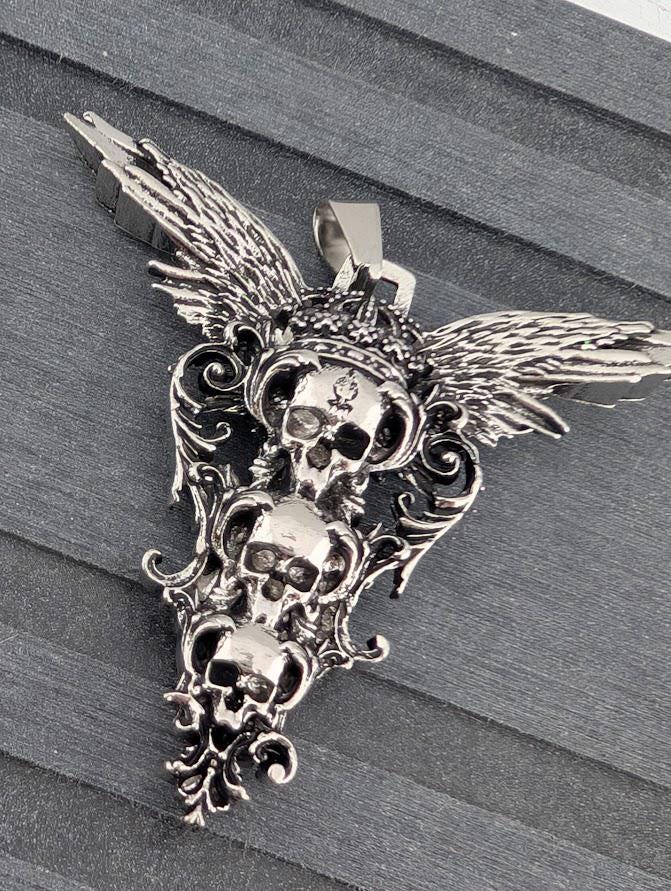 A silver-colored pendant with an intricate design featuring two skull-like figures with wings, set against a dark background.