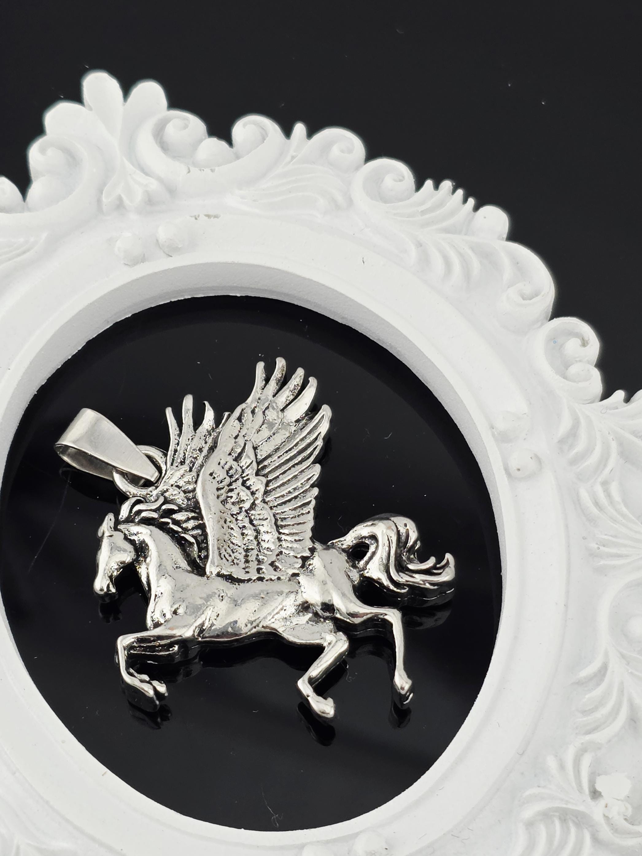 A silver pendant in the shape of a winged creature, possibly a mythical or fantastical animal, is displayed against a black background within an ornate white frame.
