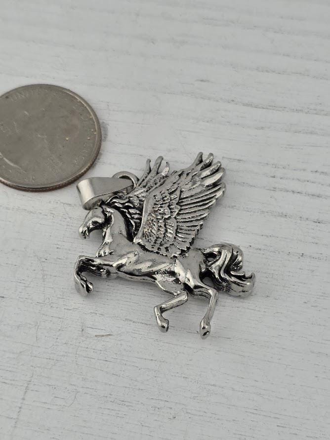 A silver-colored pendant in the shape of a winged horse, with a small coin placed next to it on a white surface.