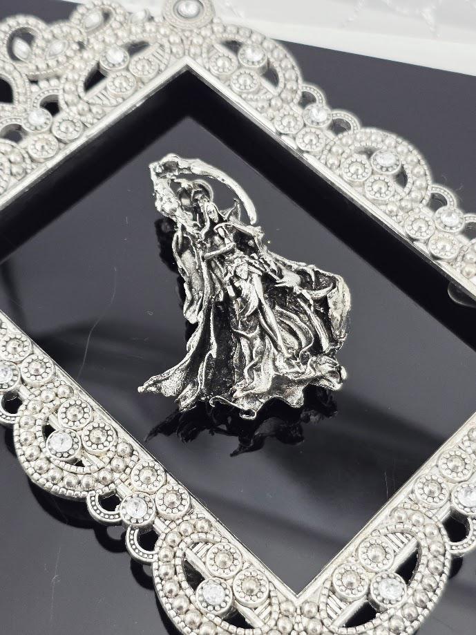 a silver-colored metal sculpture or brooch with a flowing, draped design, placed within an ornate, diamond-encrusted frame.