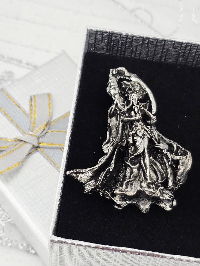 a silver-colored metal sculpture or brooch with a flowing, draped design, placed within an ornate, diamond-encrusted frame.