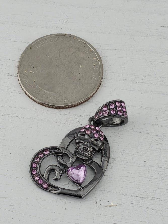 A silver pendant with a heart-shaped design and a skull-shaped design, adorned with pink gemstones, is displayed against a black background within a white ornate frame.