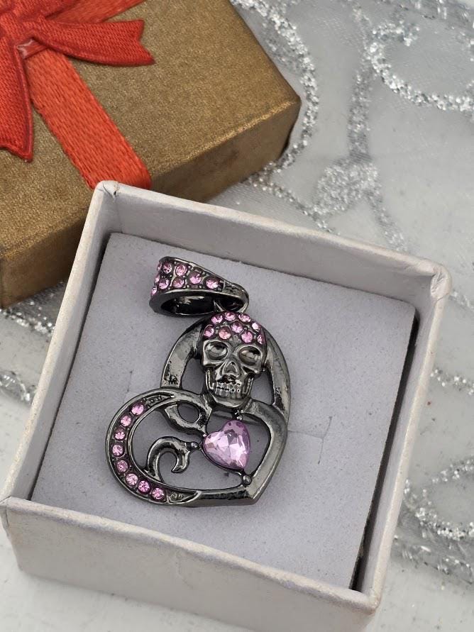 A silver pendant with a skull design and a heart-shaped gemstone is displayed in a white jewelry box.