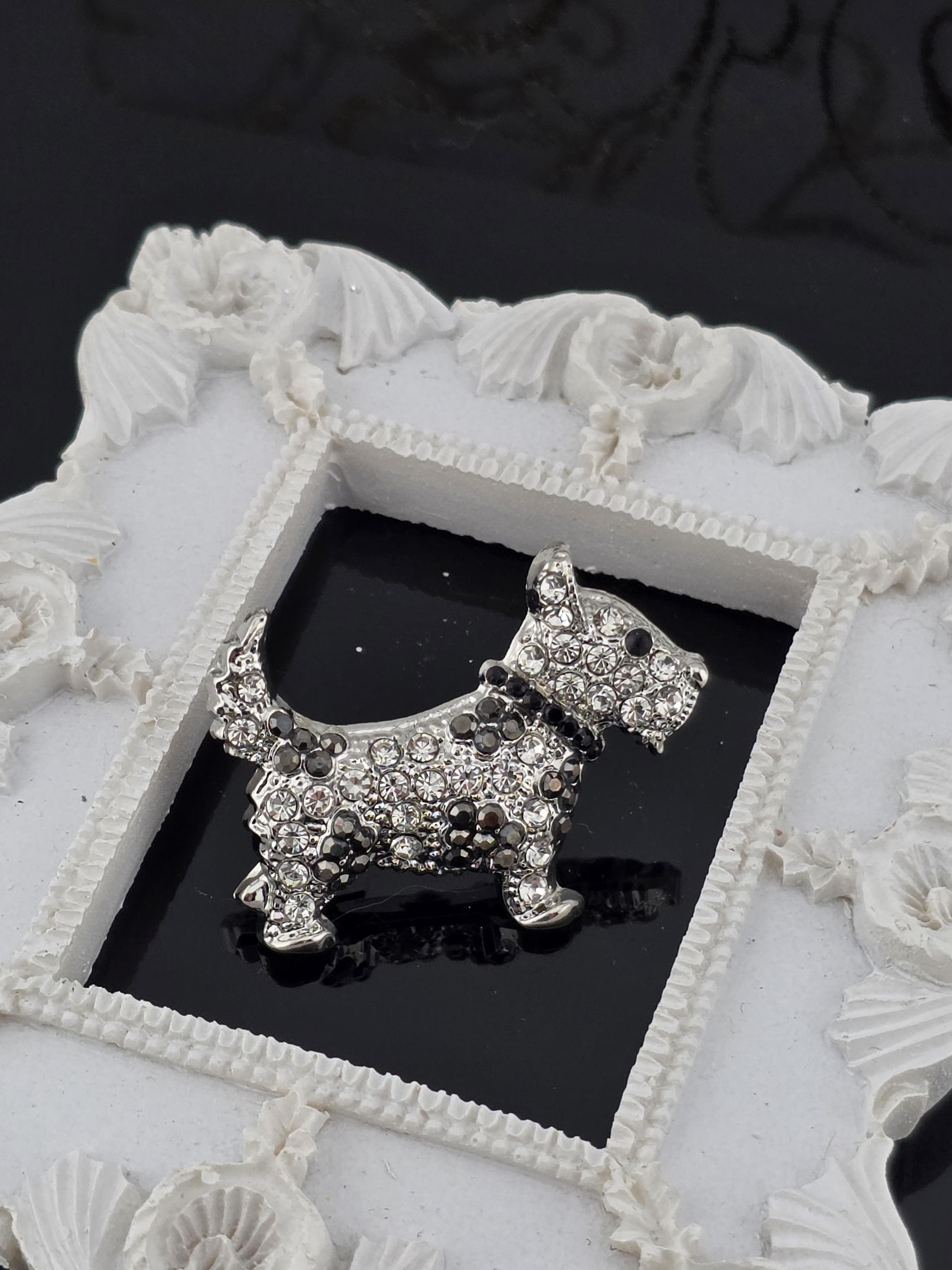 A close-up of a decorative white frame with a silver-colored ornamental piece, possibly a brooch or pendant, displayed within the frame.
