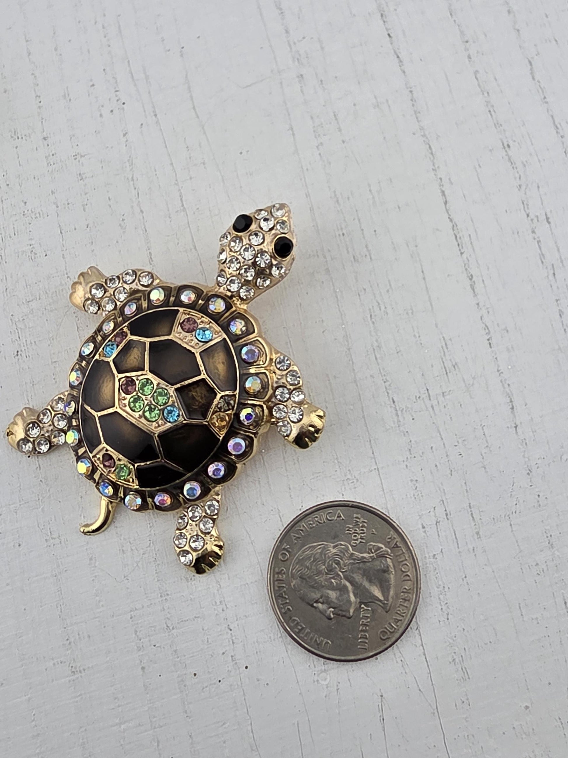 A decorative turtle brooch with rhinestones and a shiny black background, placed next to a United States quarter.