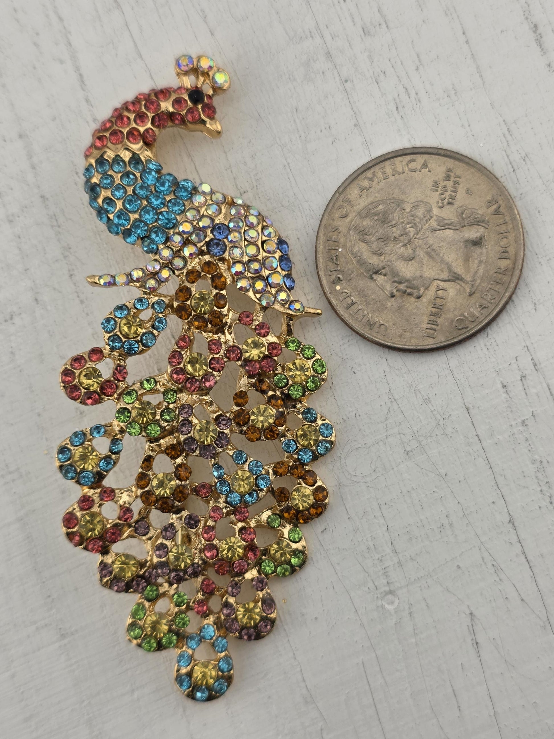 A colorful, sparkling rhinestone brooch is placed next to a United States quarter coin on a white surface.