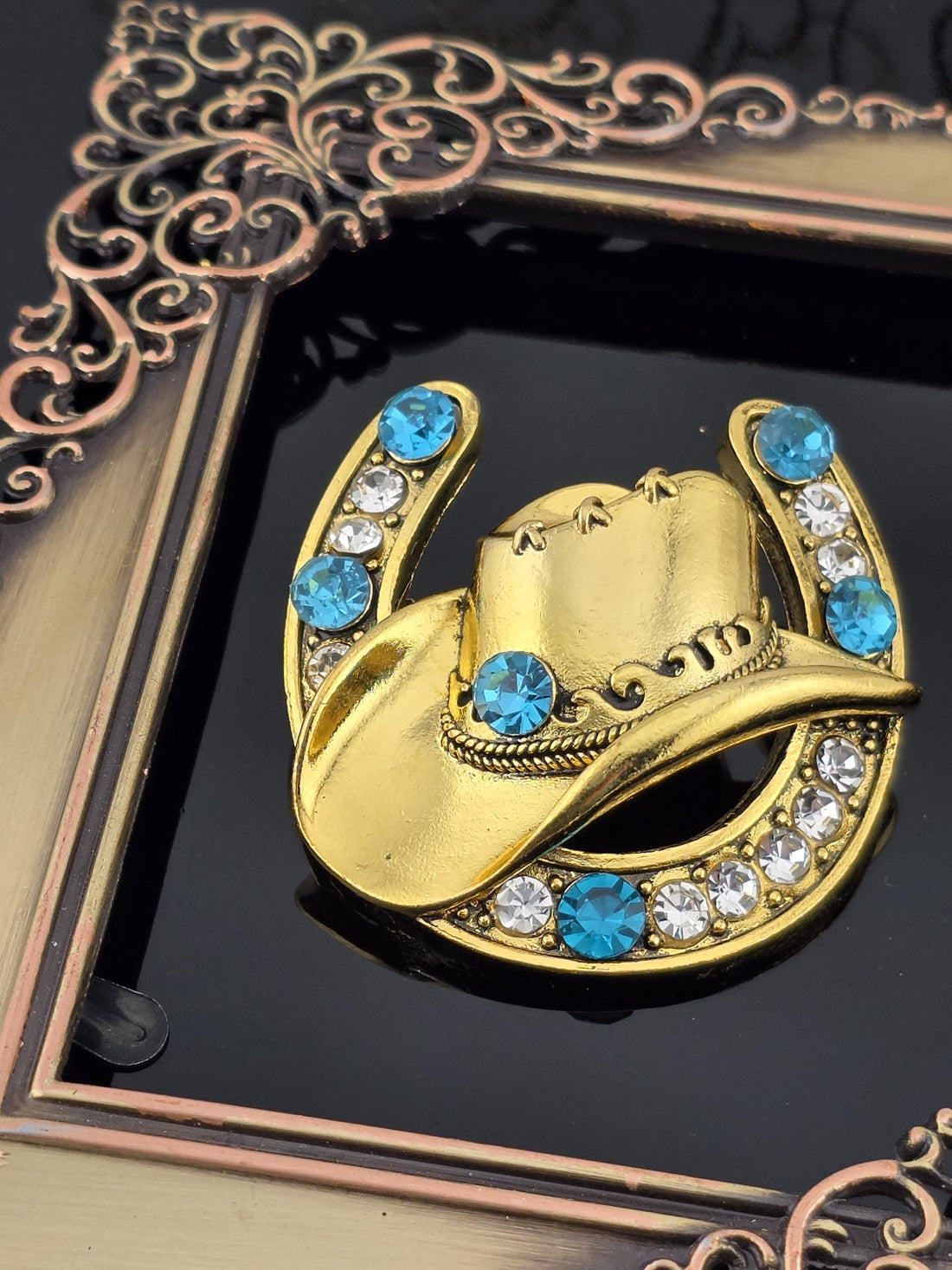 A decorative gold brooch with a blue gemstone and white gemstone accents, placed on a black background within an ornate frame.