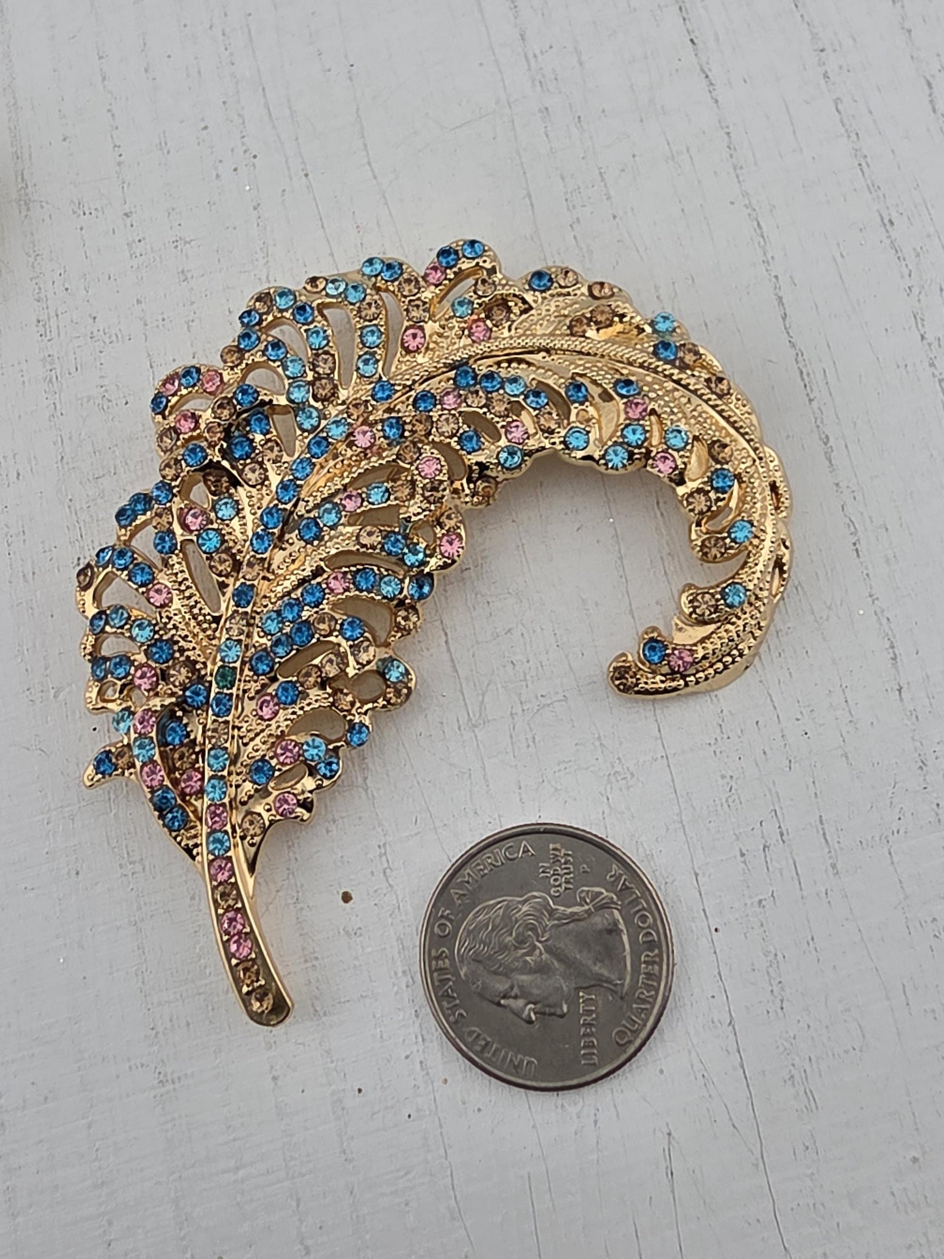a decorative gold brooch with a feather-like design, adorned with blue and pink gemstones. The brooch is placed next to a coin, which appears to be a United States quarter.