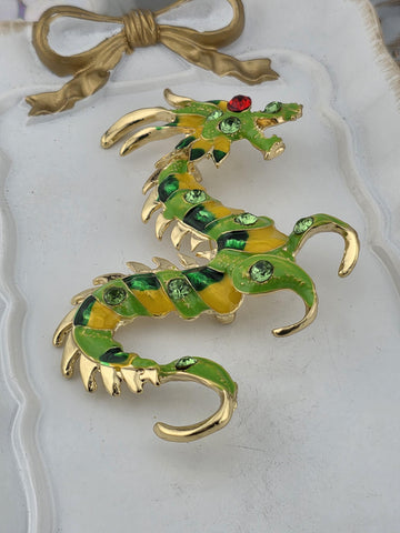A decorative dragon brooch with green and yellow scales and a red gemstone on its head, displayed on a white surface.