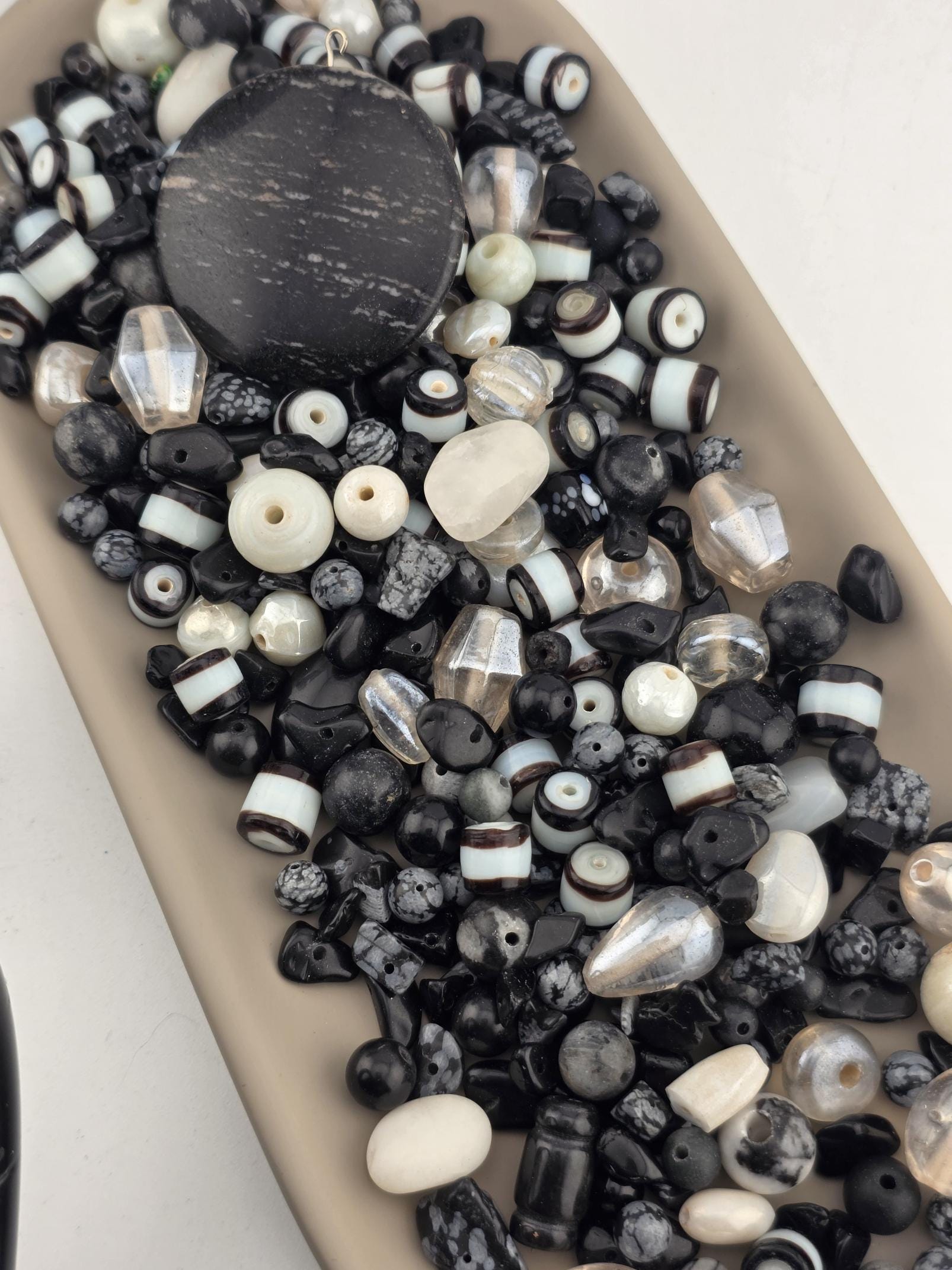 A collection of various beads in shades of black, white, and gray, including glass beads, stone beads, and metal beads, arranged on a surface.