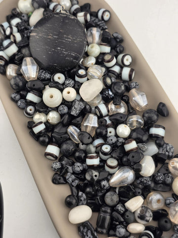 A collection of various beads in shades of black, white, and gray, including glass beads, stone beads, and metal beads, arranged on a surface.