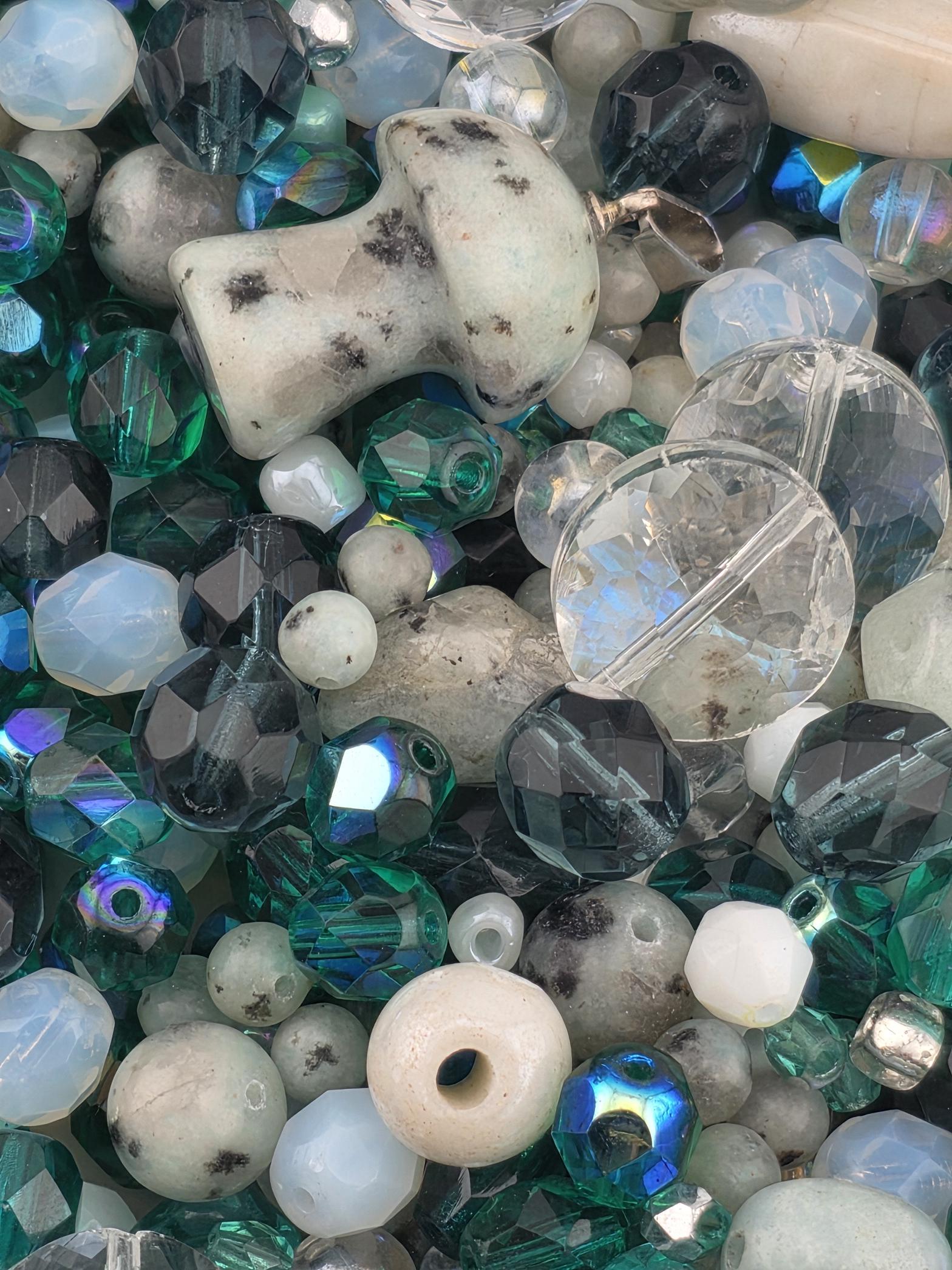 The image is a close-up of various glass beads and stones, including clear, white, blue, green, and black beads, as well as some larger stones.