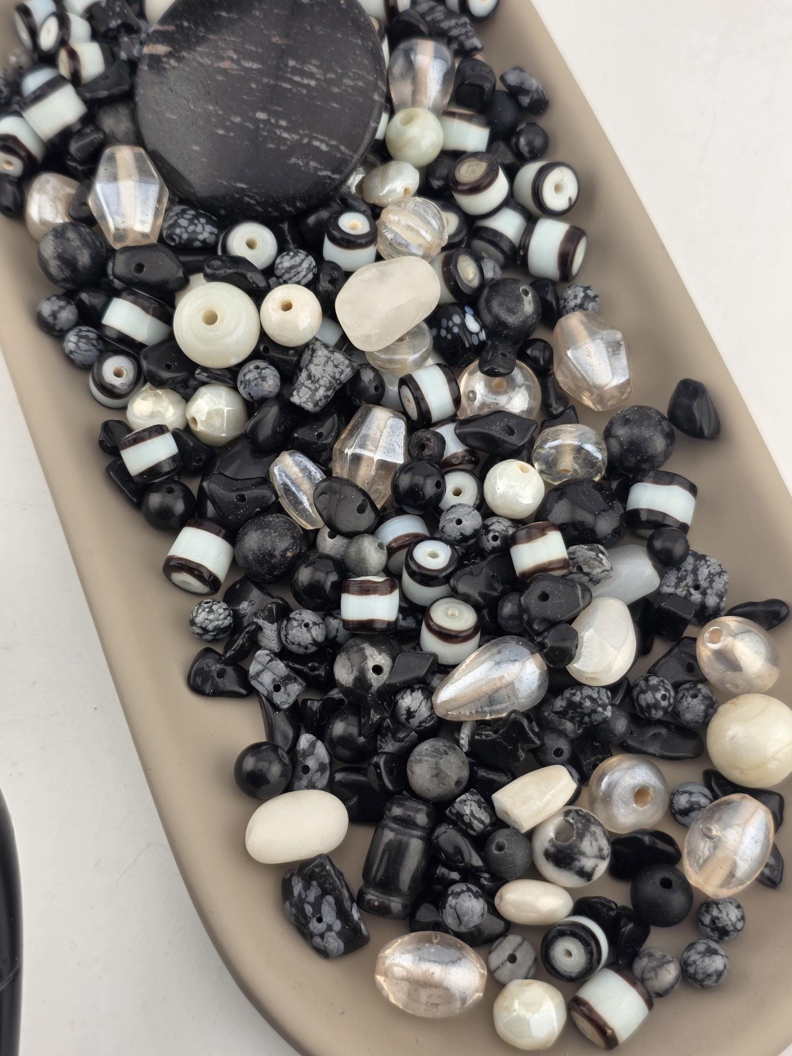 A collection of various beads in different shapes, sizes, and colors, including clear, black, white, and gray beads, arranged on a beige surface.