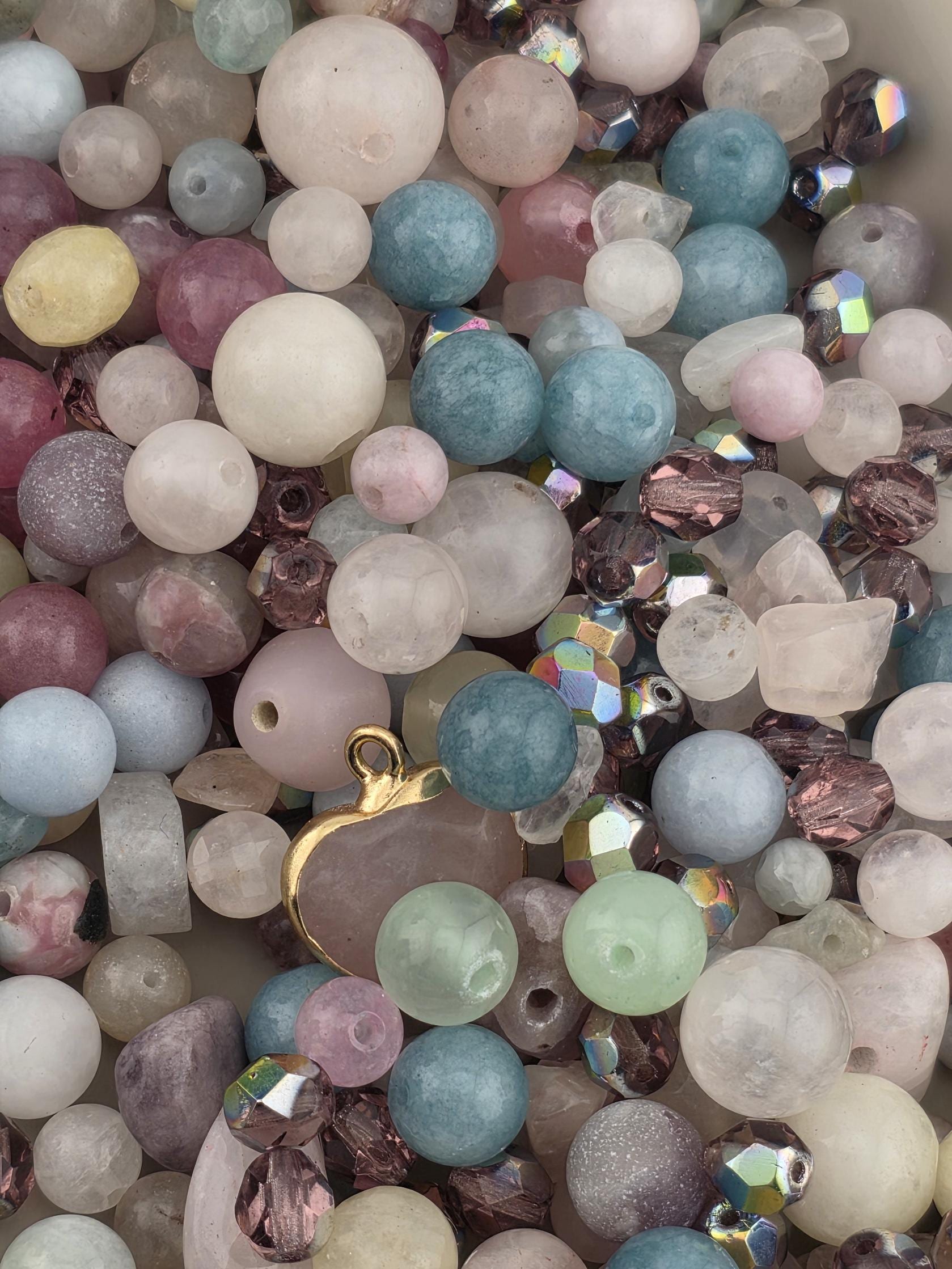 The image is a close-up of a collection of various beads and gemstones, including clear, blue, pink, and purple beads, as well as some larger gemstones.
