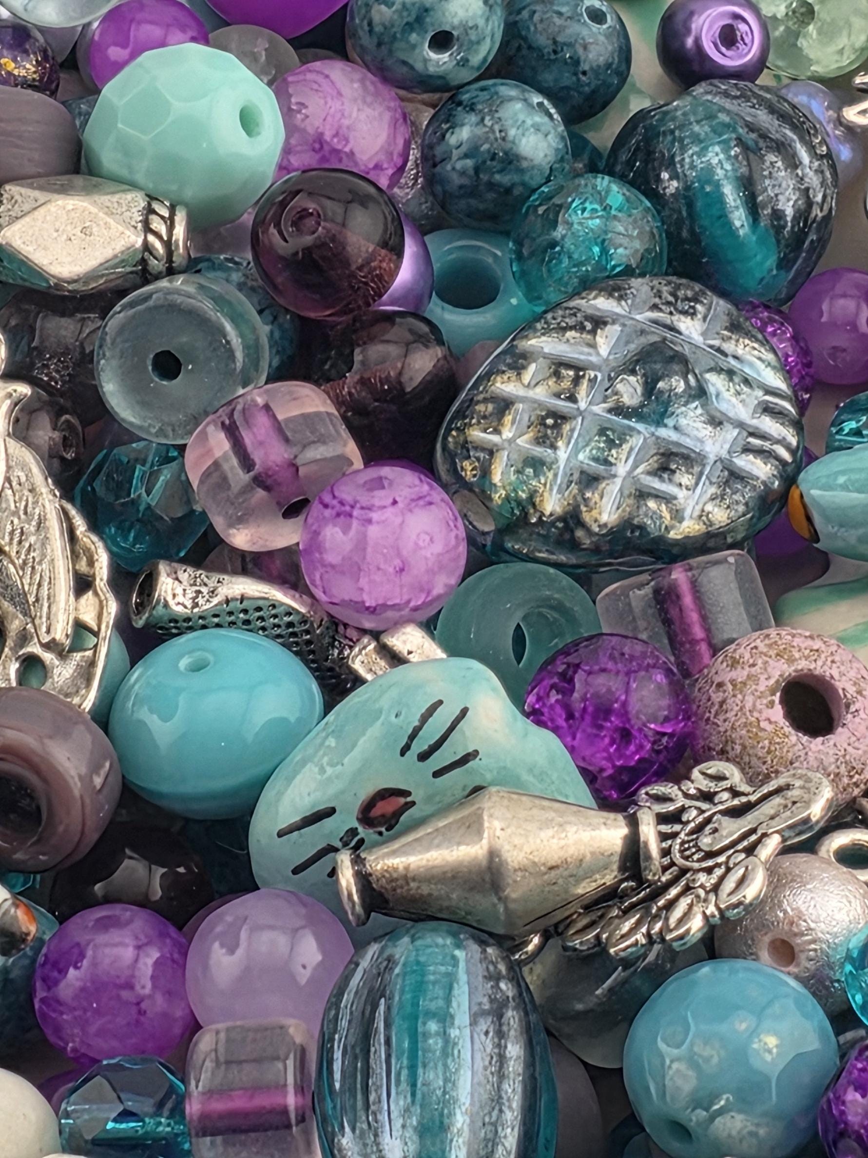 The image is a close-up of a collection of colorful beads, including glass beads in various shades of blue, purple, and green, as well as metal beads with intricate designs.