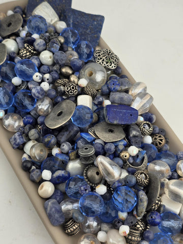 A collection of various beads, charms, and metal pieces in shades of blue, silver, and white, arranged in a visually appealing manner.