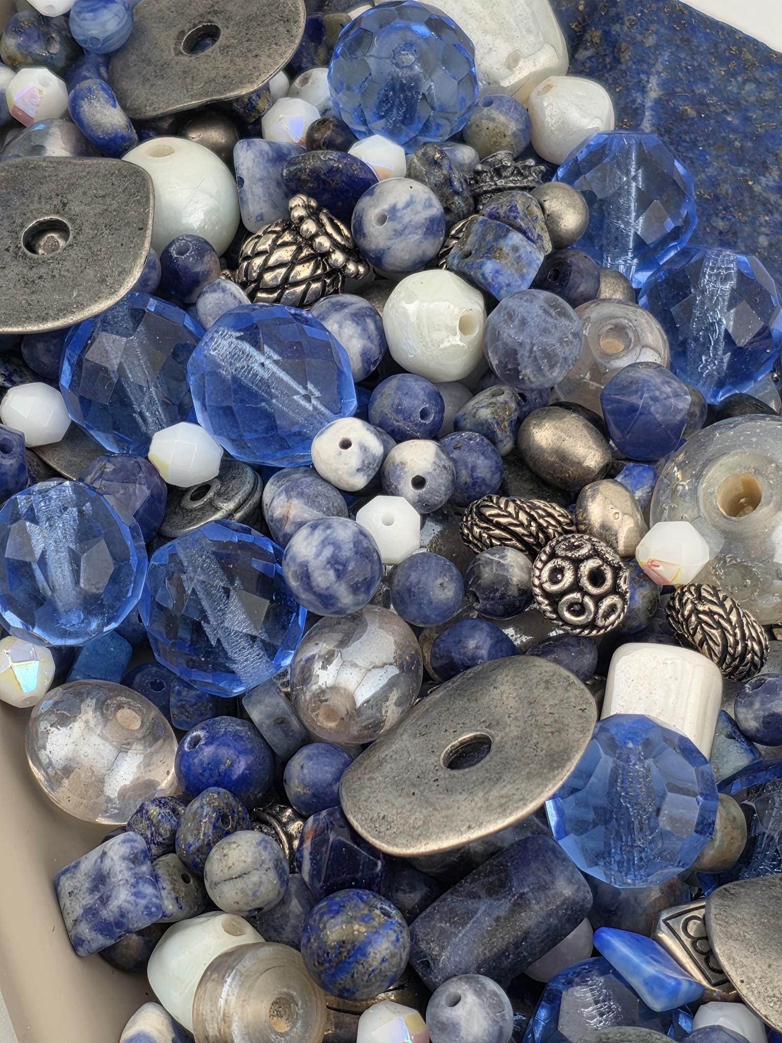 The image is a close-up of various beads and buttons in shades of blue, silver, and white, creating a visually striking and diverse collection.
