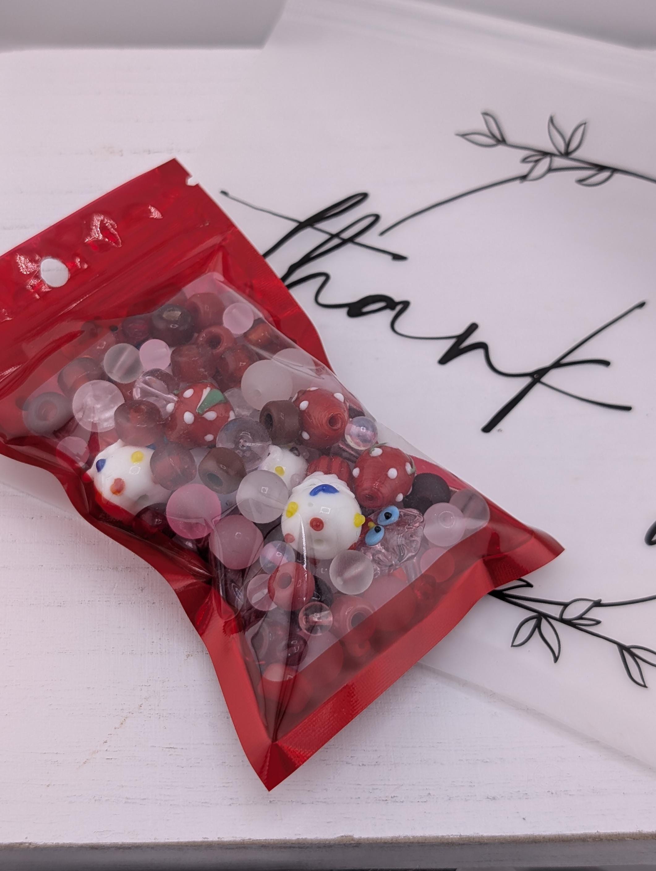A red plastic bag filled with colorful beads is placed on a white surface next to a white card with black text.