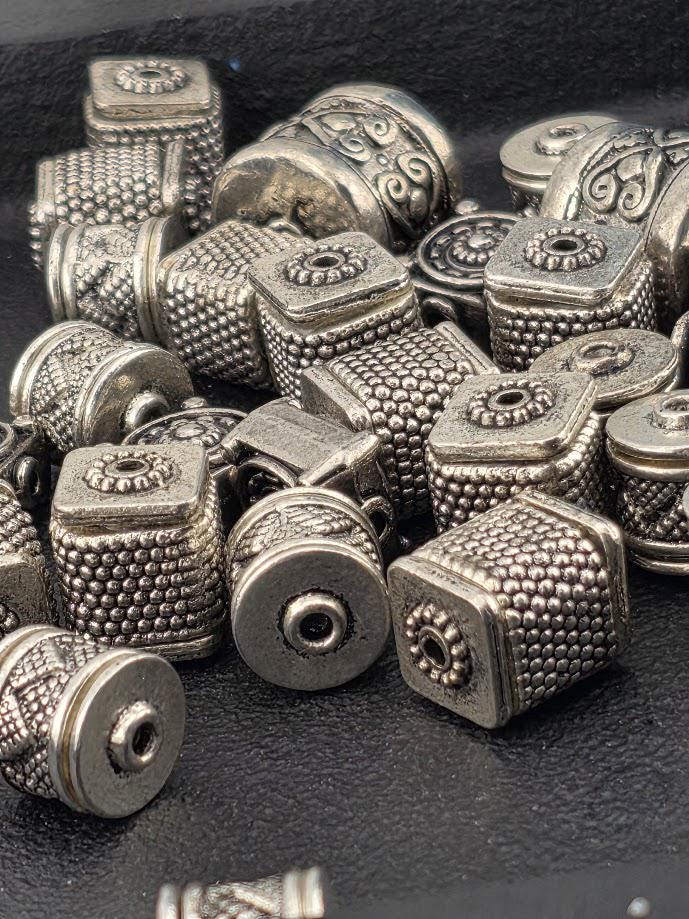a collection of various metal beads and charms, including square and round shapes, arranged on a dark surface.