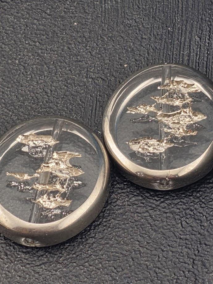 two clear glass beads with a metallic, tree-like design on them, resting on a textured surface.