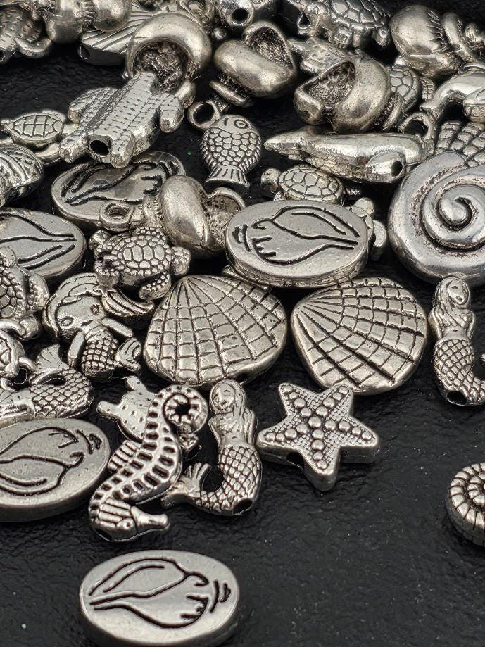 various silver-colored metal charms, including fish, shells, and other sea-themed objects, scattered on a dark surface.