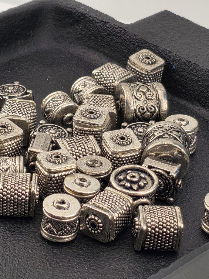 a collection of various metal beads and charms, including square, round, and ornate designs, arranged on a black surface.
