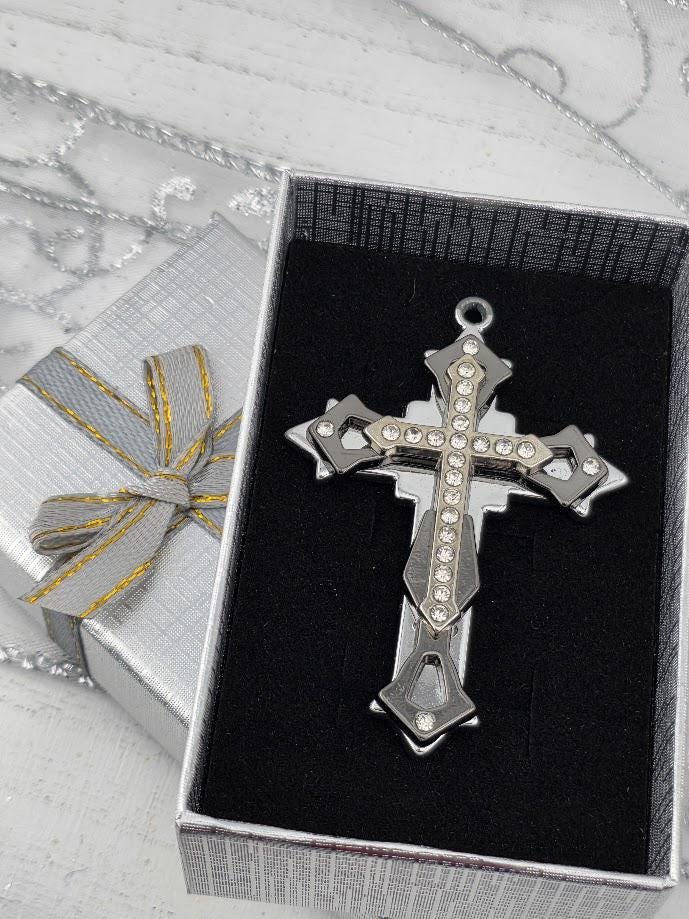 A silver cross pendant with rhinestones is displayed in a silver box with a gold ribbon bow.